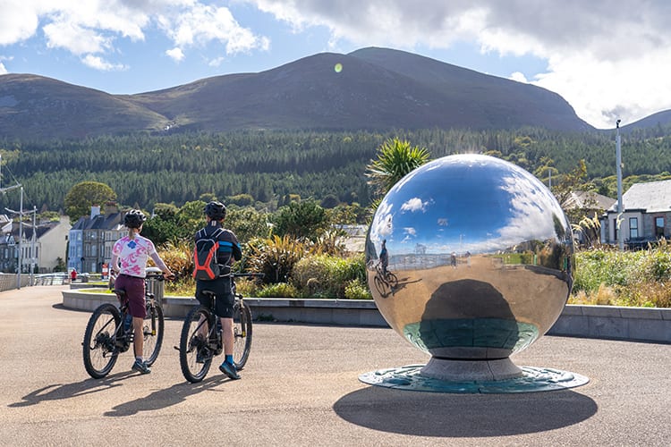 Two cyclists beside the giant spherical sculpture on Newcastle promenade with the mourne mountains in the background