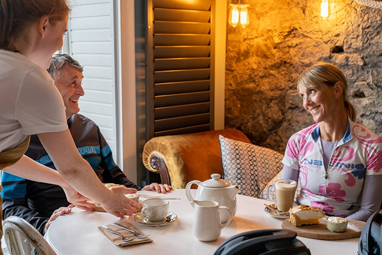 Male and female cyclists being served with hot drinks and dessert in an old stone building